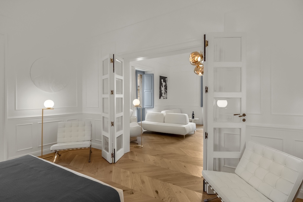 Dettaglio della porta in vetro aperta che separa il soggiorno dalla camera da letto dell'elegante suite Nora con poltrone, lampade moderne e stile artistico