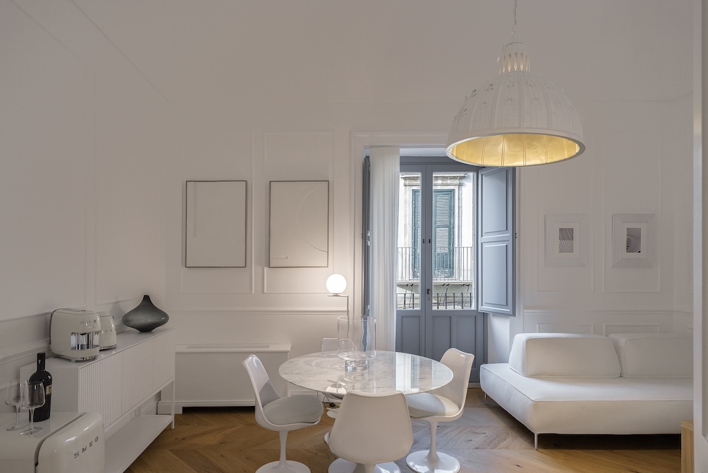 Dettaglio del soggiorno dell'elegante suite Christine con tavolo e sedie, lampade moderne, divano, macchina del caffè e bollitore e finestra con vista sulla città di Noto